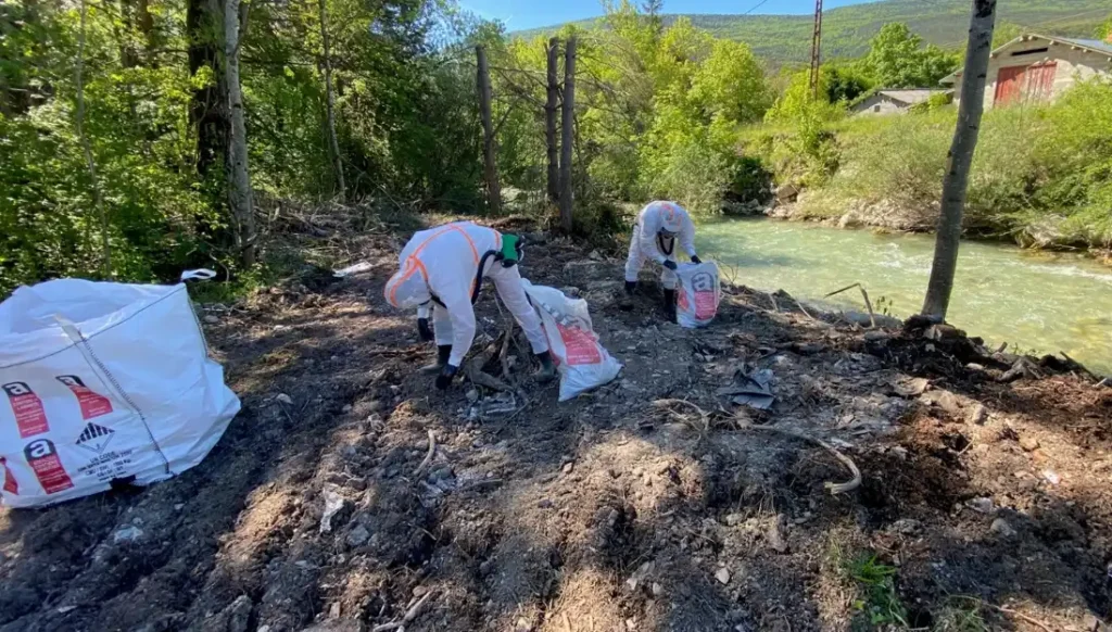 Deux personnes en combinaison blanche avec chacune un sac blanc sont en train de ramasser de l'amiante parmi les gravats du chantiers. On distingue le Buëch puis la digue et des granges en arrière plan.